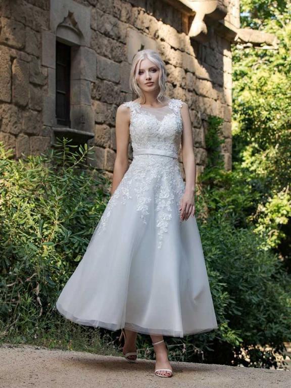 Junge Frau im weißen Hochzeitskleid vor einer alten Steinfassade und üppigem Grün.