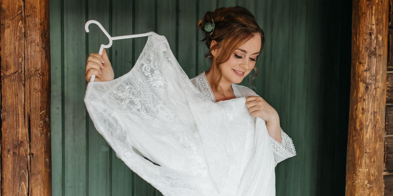 Eine junge Frau präsentiert ein großes, weißes Spitzenkleid vor einem Hintergrund mit vertikalen Holzpfosten.