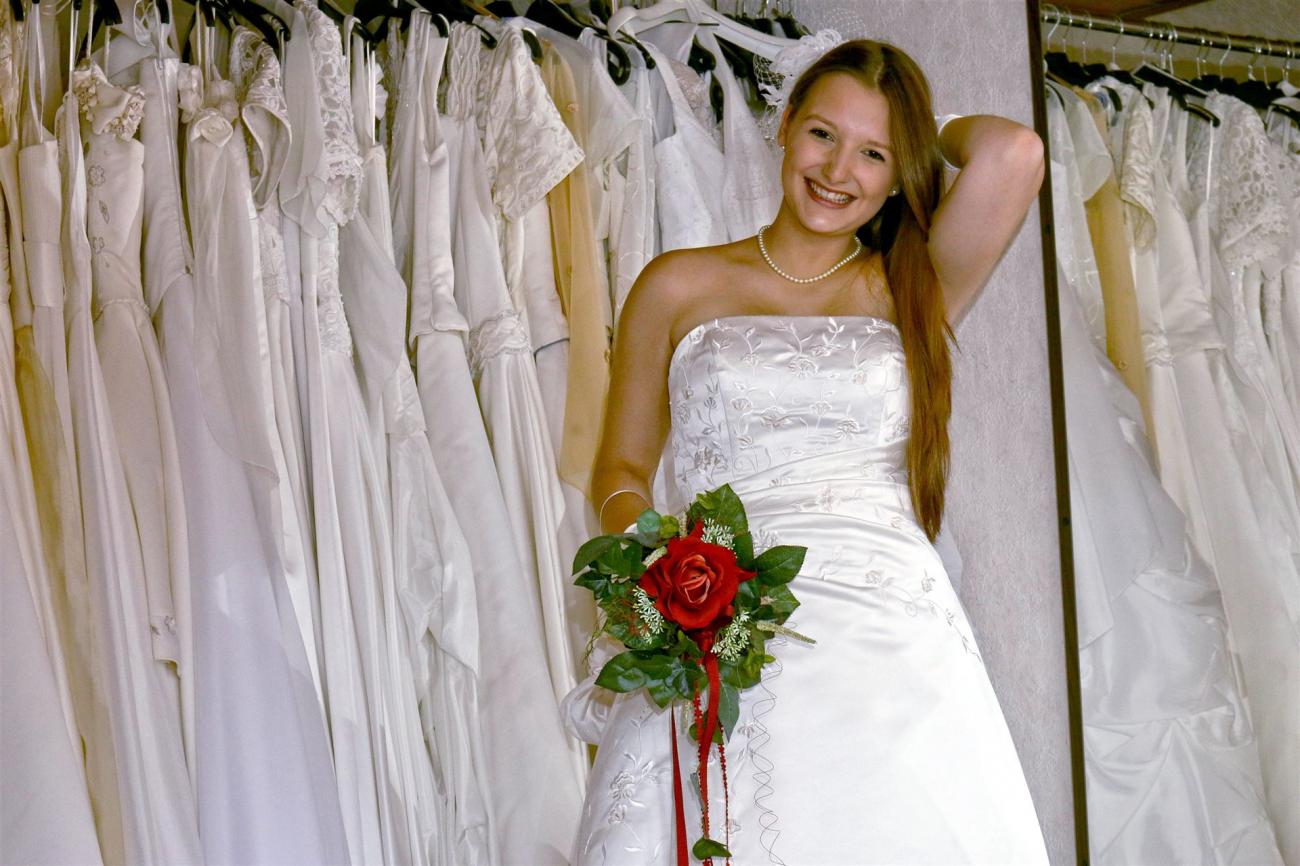 Eine lächelnde junge Frau in einem weißen Brautkleid vor einem Hintergrund voller aufgehängter Hochzeitskleider.