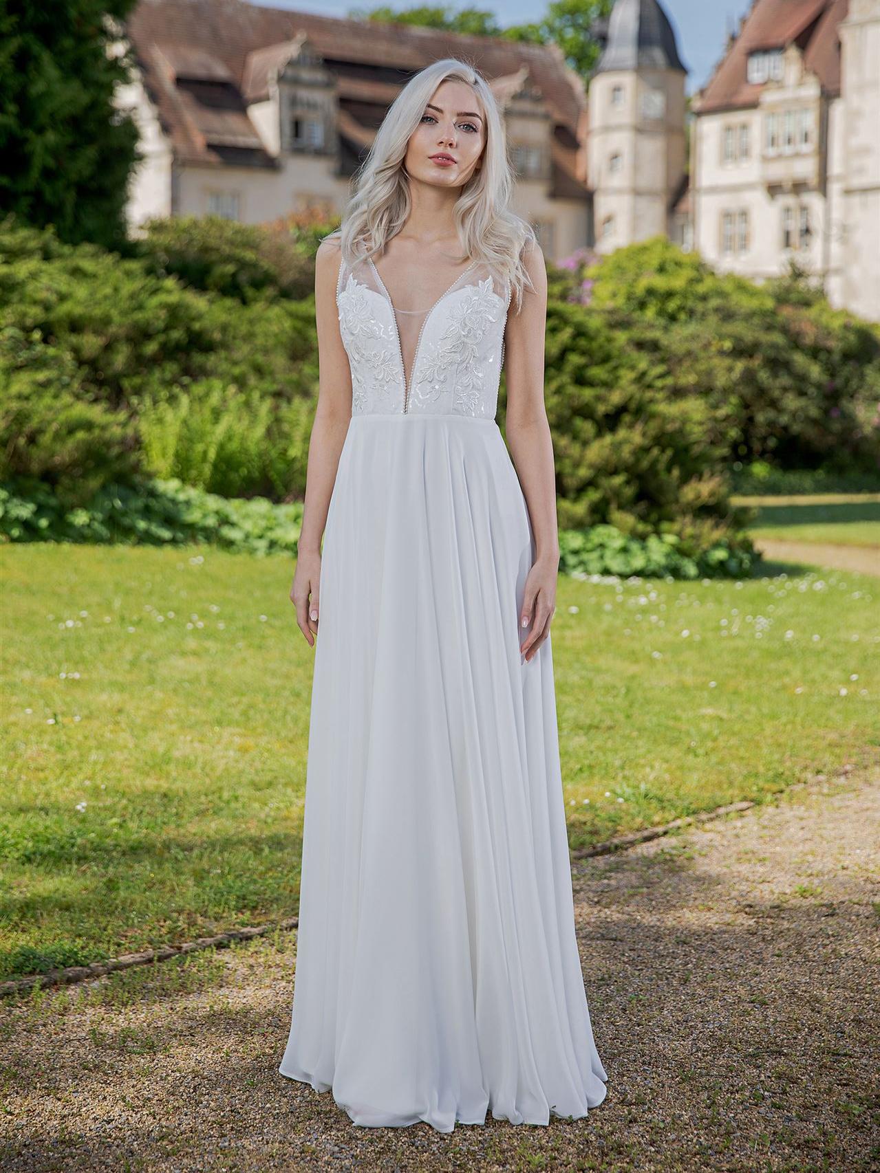 Eine Frau in einem weißen Hochzeitskleid steht auf einer Wiese vor einem historischen Schlossgebäude.