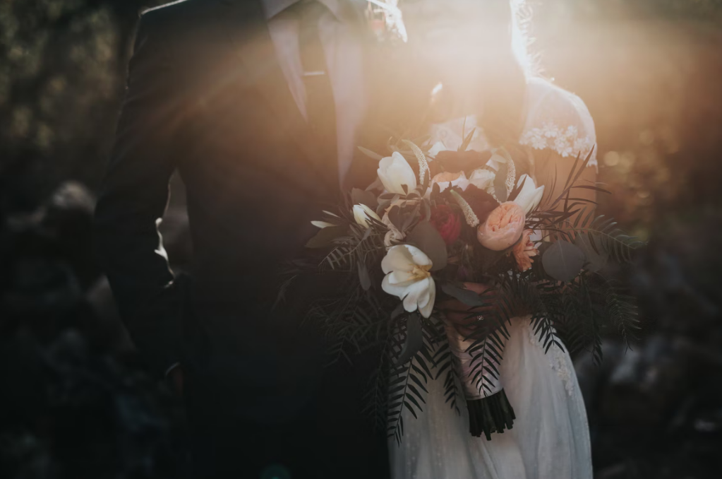 Ein Hochzeitspaar bei dramatischem Gegenlicht, wobei die Braut einen großen, blumigen Strauß hält.