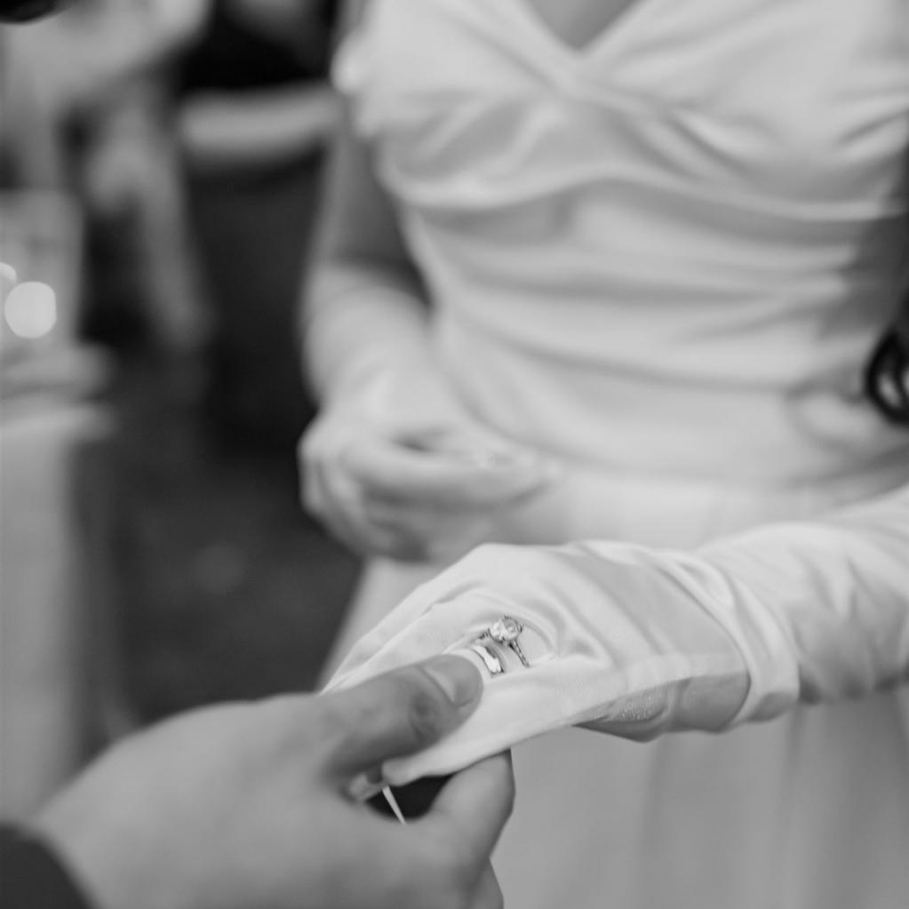 Schwarz-Weiß-Nahaufnahme von zwei Händen, deren Ringe im Mittelpunkt stehen, während eine Person ein weißes Abendkleid trägt.