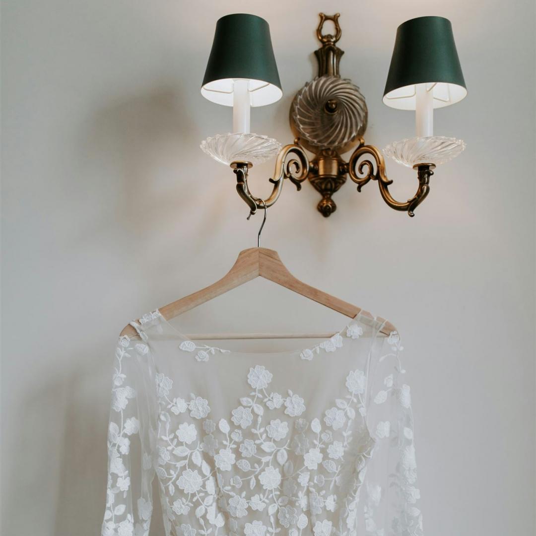 Ein weißes Spitzenkleid mit Blumenapplikationen hängt vor einer dekorativen Wandleuchtanordnung.