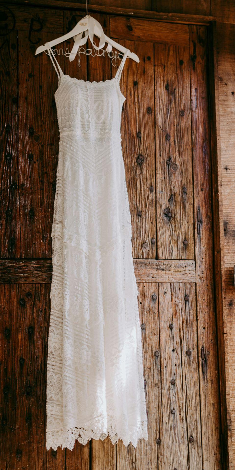Ein langes, weißes Spitzenkleid hängt vor einer rustikalen Holztür.