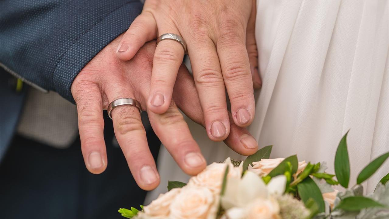 Close-up von zwei Händen, die sich bei einer Hochzeitszeremonie berühren, vor einem weißen Kleid und einem Blumenstrauß.