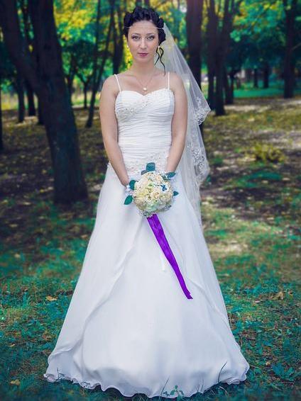 Eine Braut in einem weißen Kleid steht in einem üppär grünen Park mit hohen Bäumen.