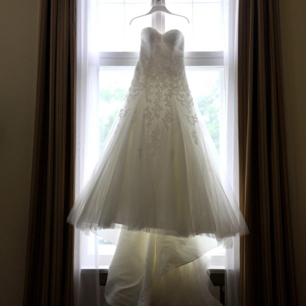 Ein weißes Brautkleid hängt vor einem großen Fenster und wird von natürlichem Licht beleuchtet.