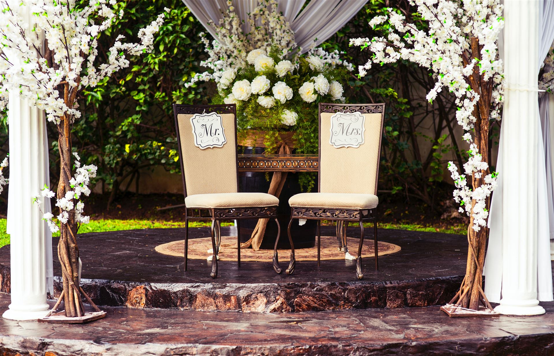 Ein romantisches Hochzeits-Setup mit zwei Stühlen für Mr. und Mrs. vor einem Blumenarrangement, eingerahmt von blühenden Bäumen und weißen Säulen.