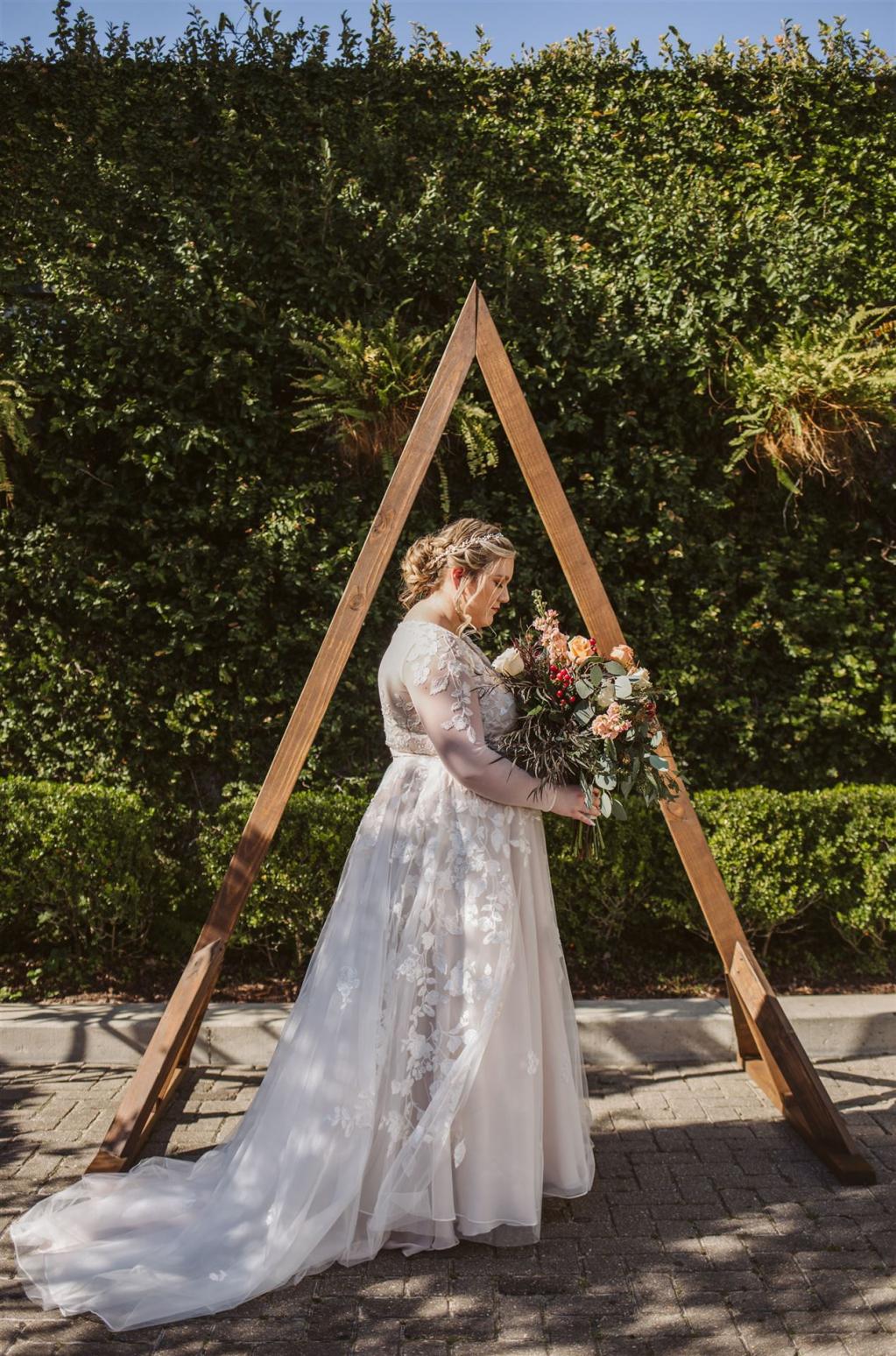 Eine Braut in einem weißen Kleid vor einem hölzernen A-Frame-Dreieck mit grünem Hintergrund.