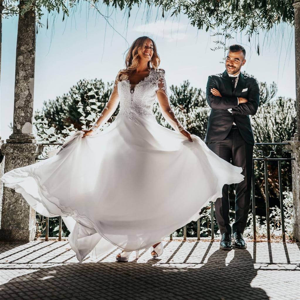 Eine fröhliche Braut in einem weißen Hochzeitskleid und ihr Bräutigam stehen unter einem begrünten Laubdach.