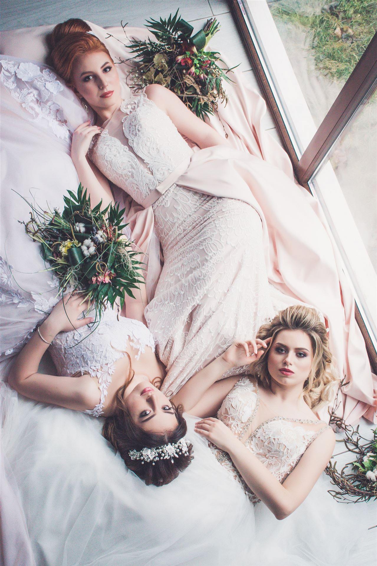 Drei Frauen posieren glamourös auf einem Bett, umgeben von fließenden, hellrosa Tüchern und grüner Dekoration.