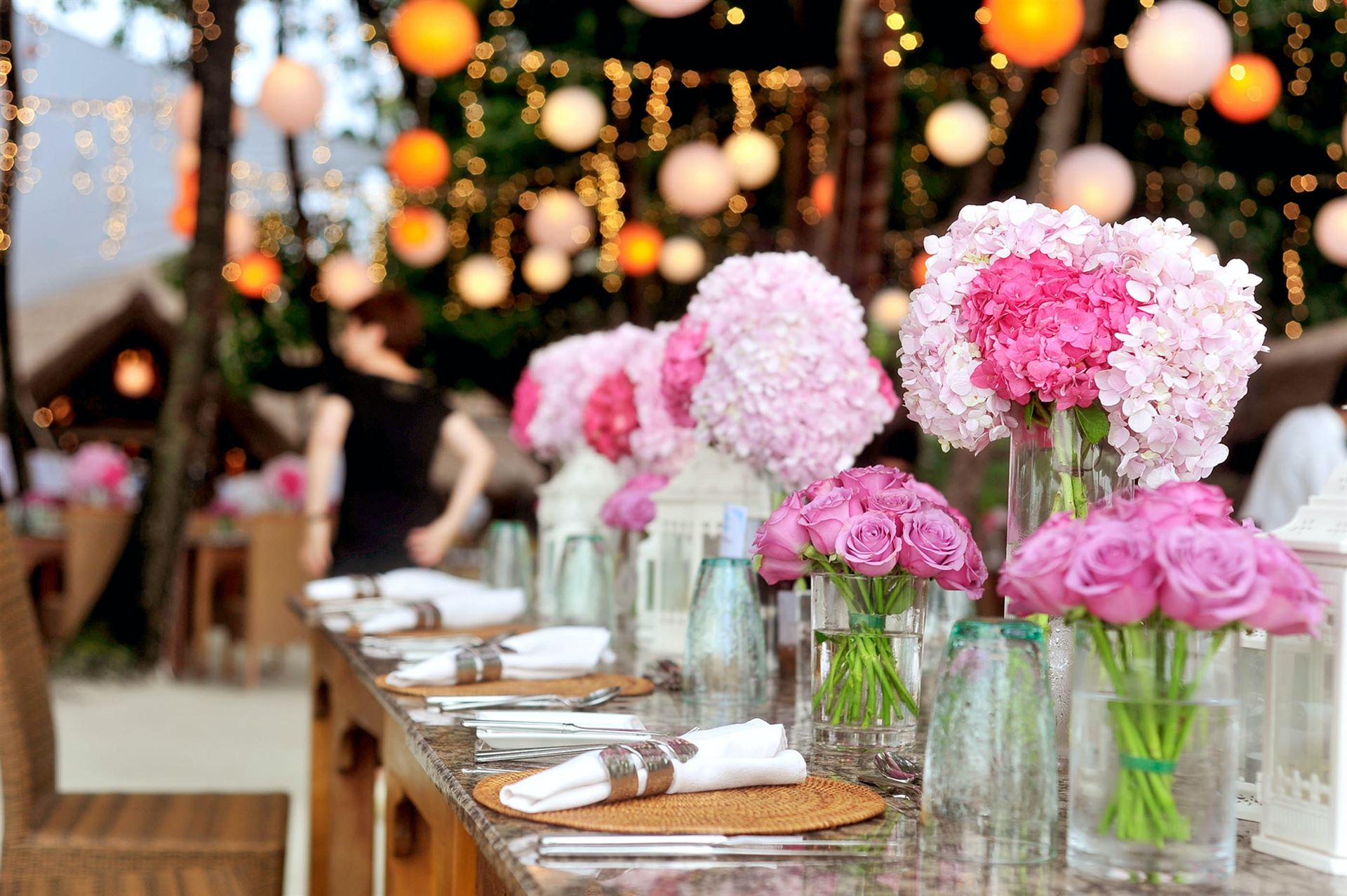 Langer Tisch, festlich dekoriert mit Blumenbouquets in verschiedenen Rosatönen und beleuchtet von warmen Lichterketten.