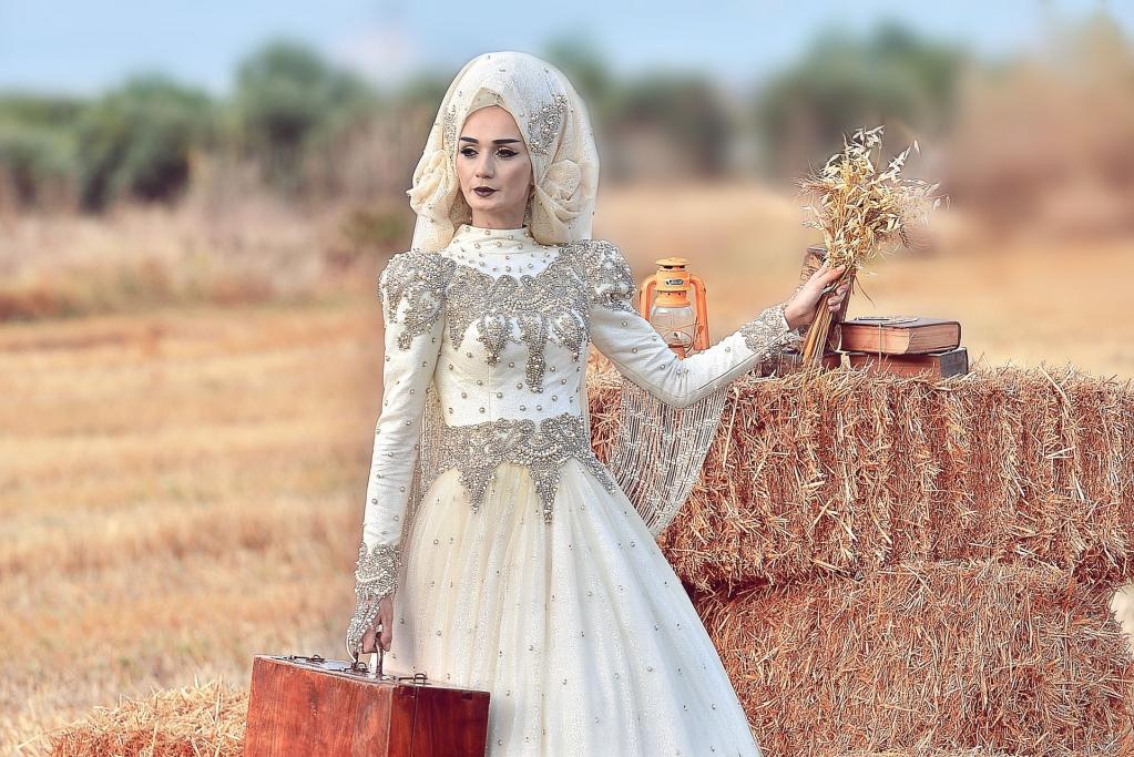 Eine Frau in einem verzierten weißen Kleid und Schleier steht auf einem Feld neben einem großen Heuballen.