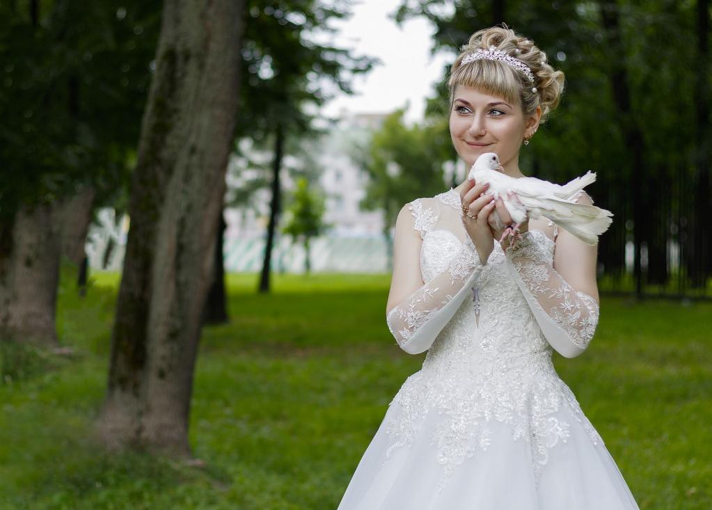 Eine junge Frau in einem weißen Brautkleid hält einen weißen Vogel in einem grünen Park.