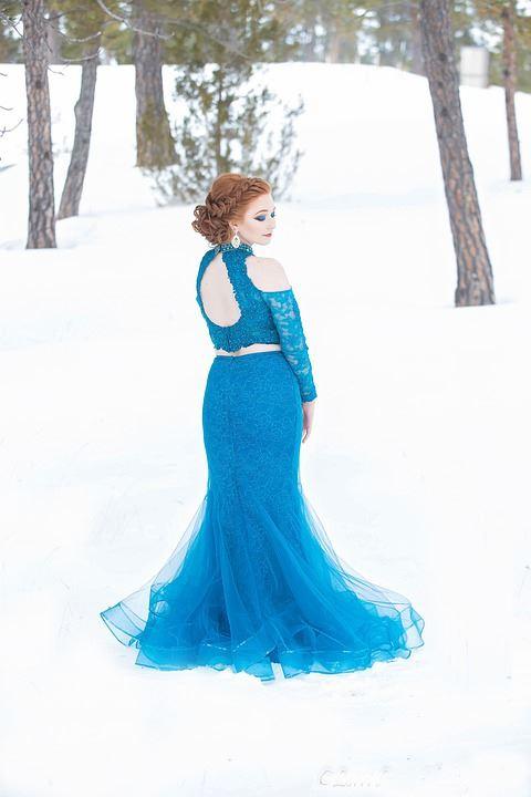 Eine Frau in einem fließenden, blauen Ballkleid steht in einer verschneiten, baumgesäumten Landschaft.
