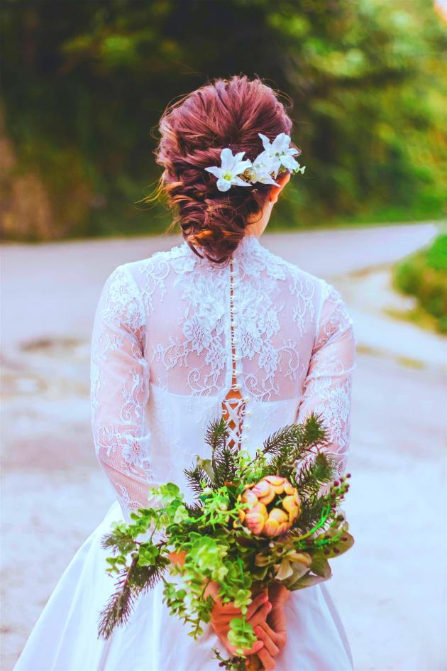 Rückenansicht einer Braut mit Blumenkranz und einem üppigen Blumenstrauß auf einem Weg.