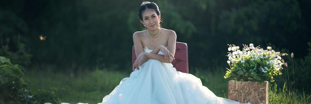 Eine Braut in einem weißen Hochzeitskleid sitzt im Grünen vor dem Hintergrund eines dunklen Waldes.