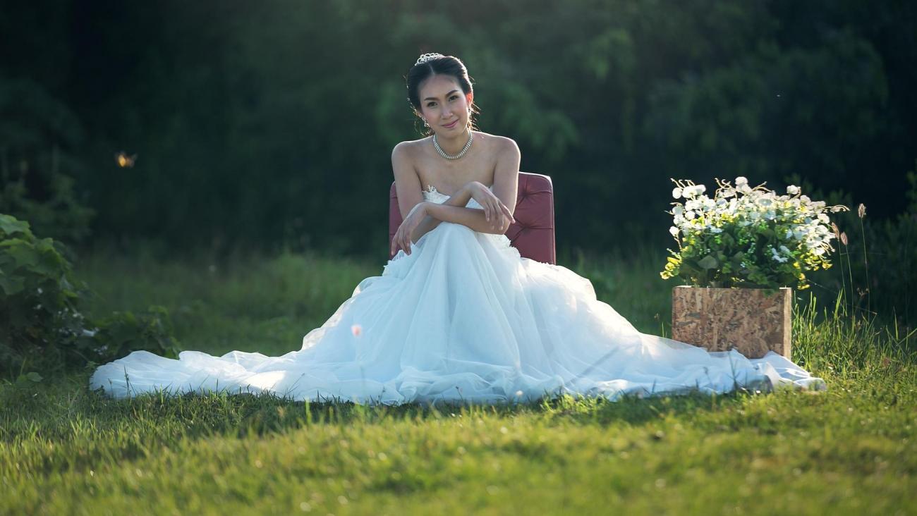 Eine Braut in einem weißen Hochzeitskleid sitzt im Grünen vor dem Hintergrund eines dunklen Waldes.