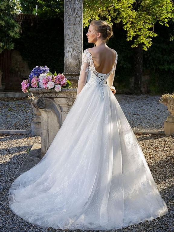 Eine Braut in einem voluminösen, weißen Hochzeitskleid steht in einem grünen Garten vor einem steinernen Sockel.