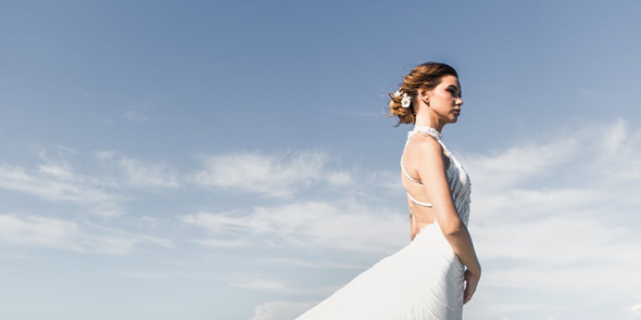 Eine Frau in einem weißen Kleid steht im Freien vor einem weiten, wolkenverhangenen blauem Himmel.