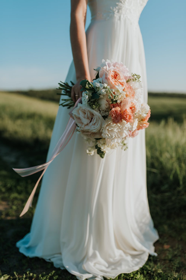 Eine Braut in einem fließenden weißen Kleid steht in einem weiten, sonnigen Feld und hält einen großen Blumenstrauß.