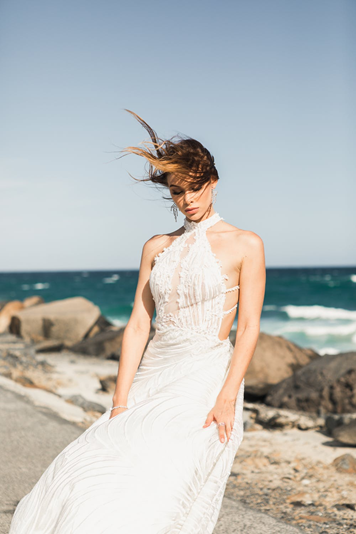 Eine Frau in einem weißen, fließenden Kleid steht an einem felsigen Strand mit Blick auf das offene Meer.