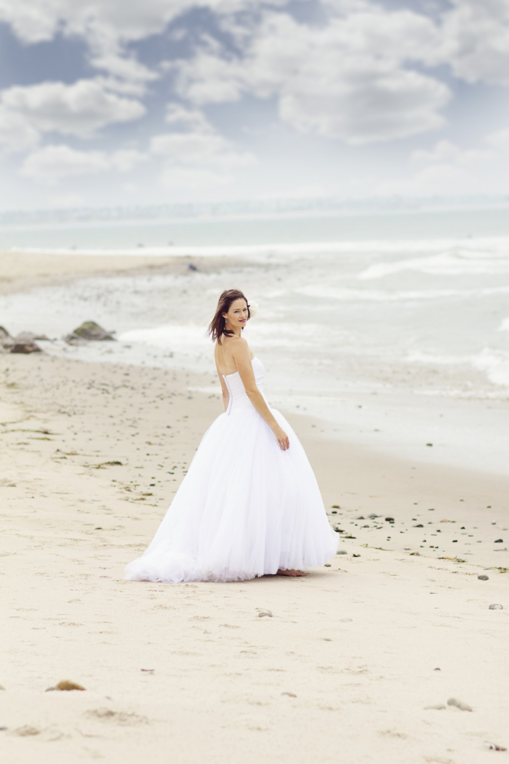 Eine junge Frau in einem weißen Hochzeitskleid steht auf einem weiten, sandigen Strand vor dem Meer.