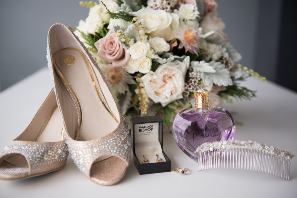 Ein Paar silberne High Heels, ein großer Blumenstrauß, eine Flasche lila Parfum und ein Kamm sind auf einer weißen Oberfläche drapiert.