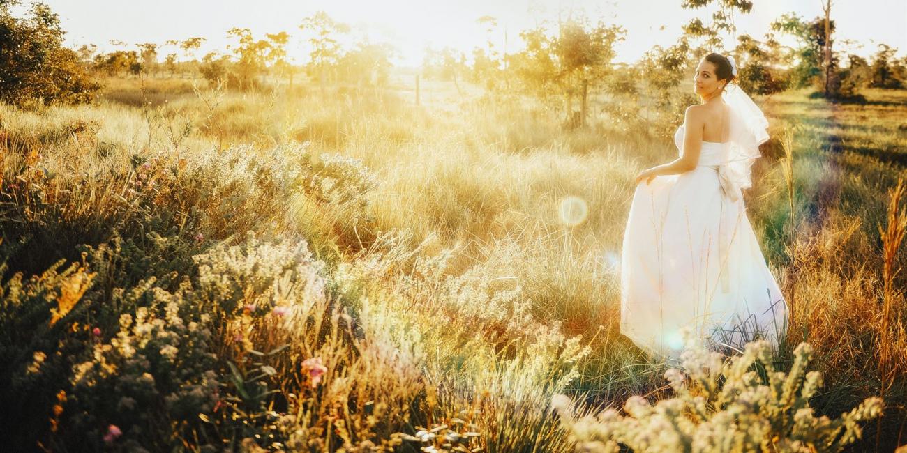 Eine Frau in einem weißen Kleid steht in einer weiten, goldgelben Wiese, umgeben von hohen Gräsern und Wildblumen bei Sonnenuntergang.