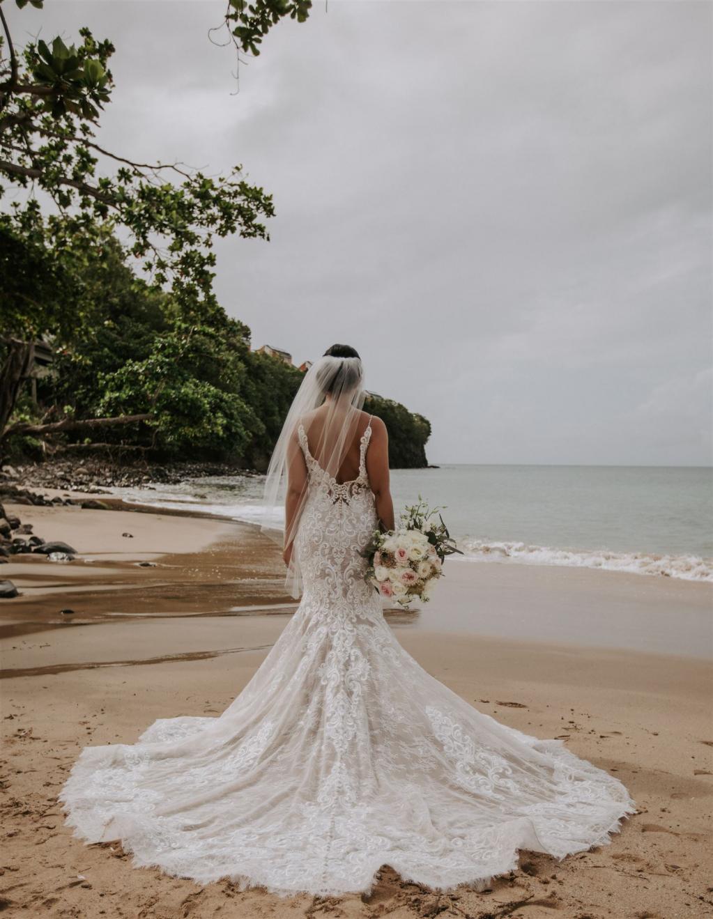 Eine Braut in einem langen, weißen Kleid steht an einem tropischen Strand und blickt auf das Meer hinaus.