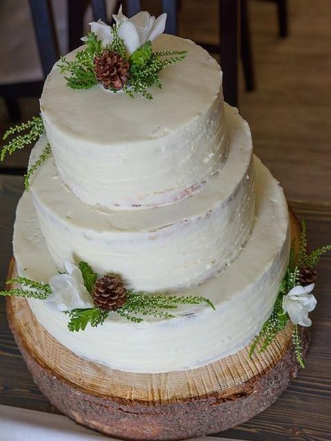 Ein dreistöckiger, rustikaler Hochzeitsturm aus weißem Kuchen, dekoriert mit Tannenzapfen und weißen Blumen auf einer Holzscheibe.