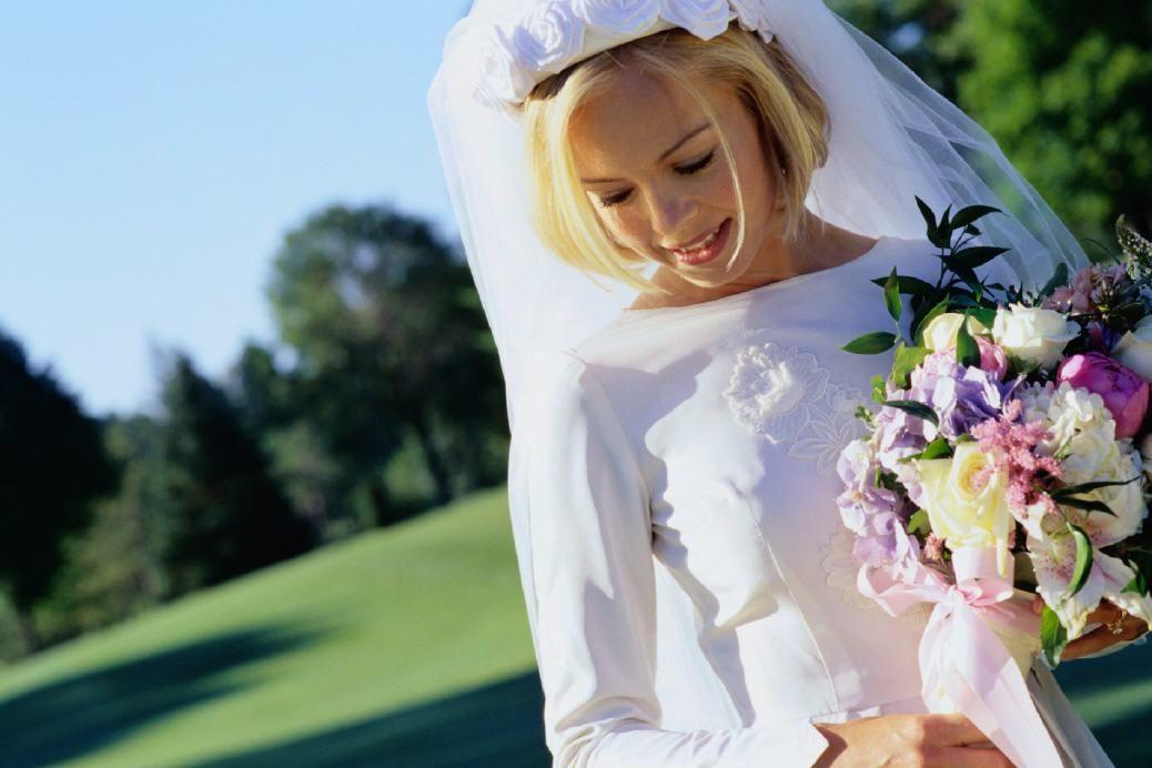 Eine blonde Braut in einem weißen Hochzeitskleid und Schleier hält einen farbenprächtigen Blumenstrauß vor einer grünen Landschaft.